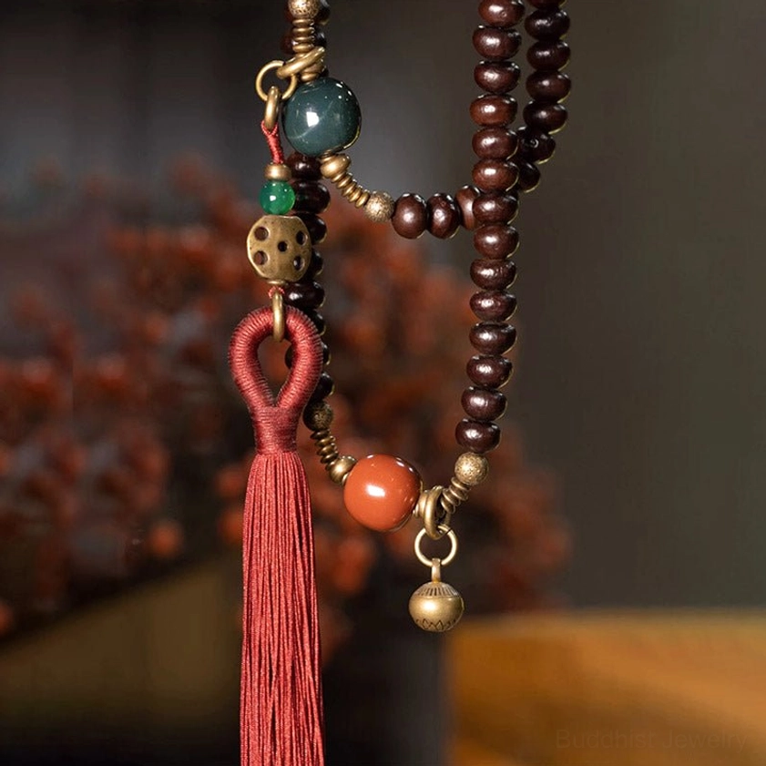 Close-up of mala tassel and brass charm details with colorful stone beads on blurred floral background