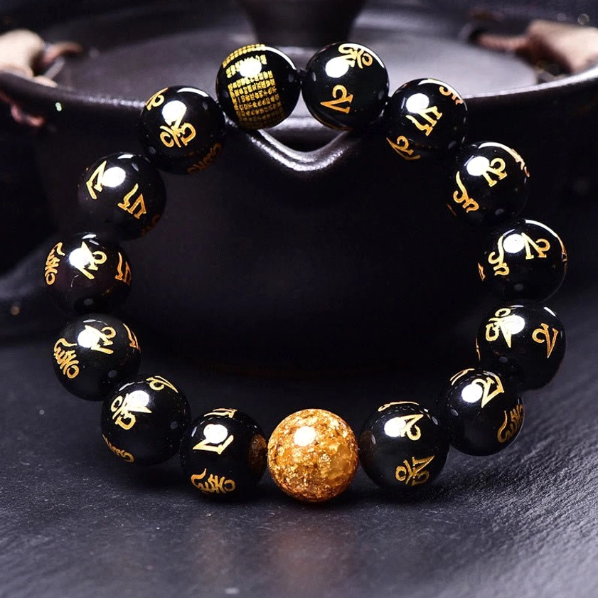Six true words Buddhist beaded bracelet top-down view showing engraved scripture bead, gold foil accent, and Sanskrit-inscribed black obsidian beads in ceramic bowl