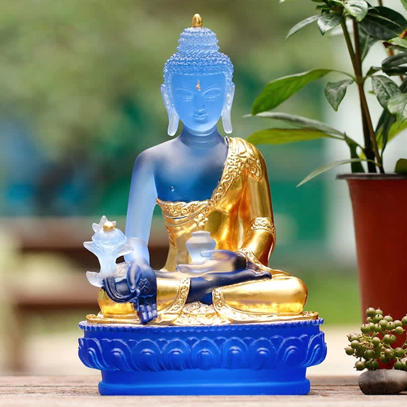 Handmade Medicine Buddha glass sculpture with frosted light blue body and gilded robe displayed outdoors on wooden table with plants