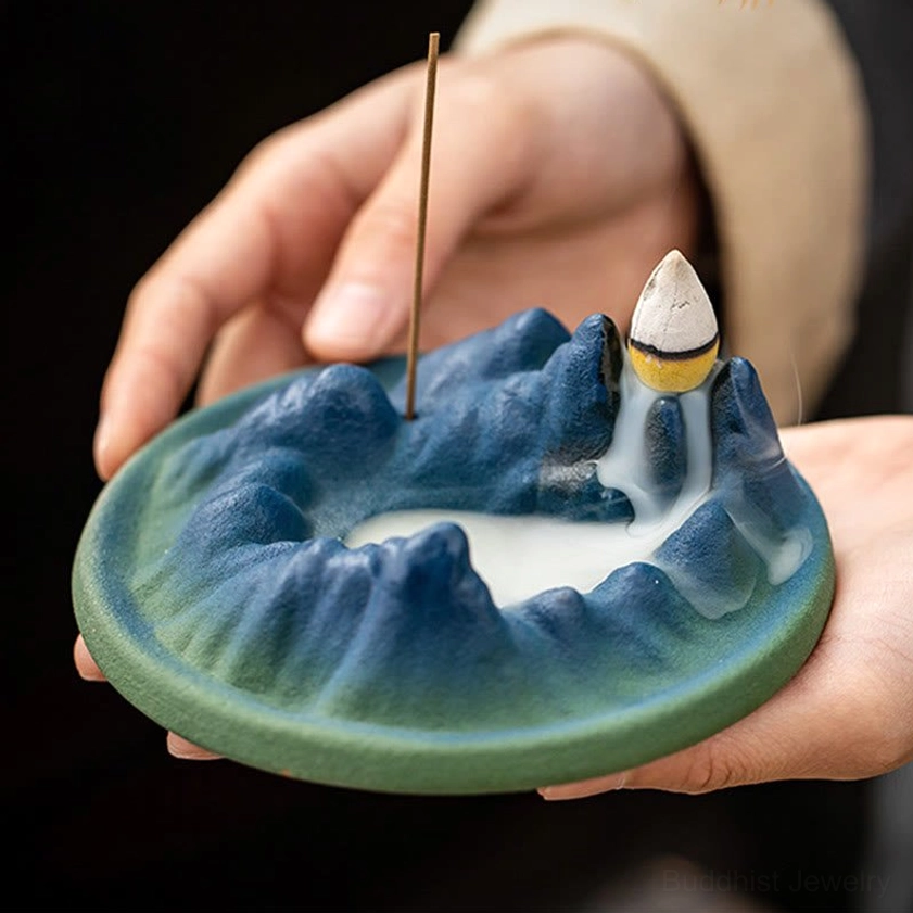 Buddhist incense burner held in open palms with both incense stick and cone installed, showing active backflow smoke and matte ceramic finish