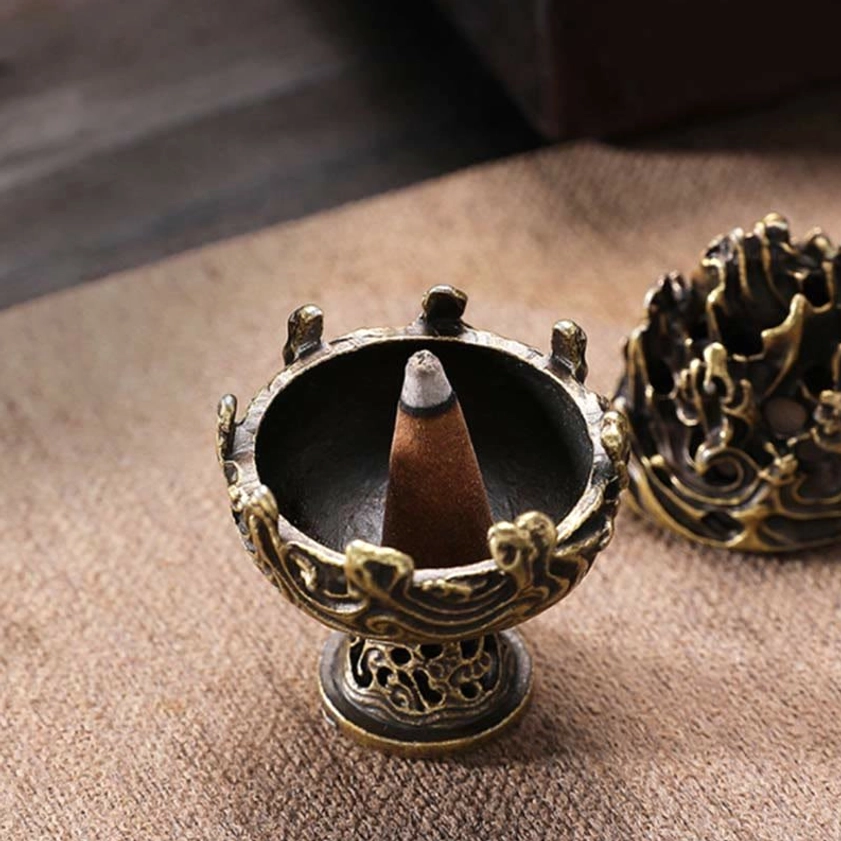 Bronze Buddhist incense burner flat lay with cone incense inside, showing carved scroll motifs and antique finish on linen cloth