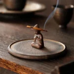 Handcrafted ceramic monk incense burner with burning sandalwood incense on wooden tea table, showing rising fragrant smoke for meditation and spiritual practice