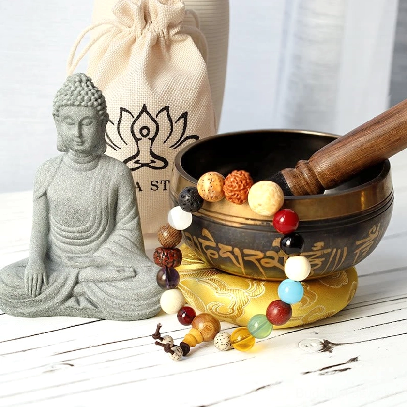 Buddhist singing bowl with mala beads and sandstone Buddha statue displayed on white wooden surface