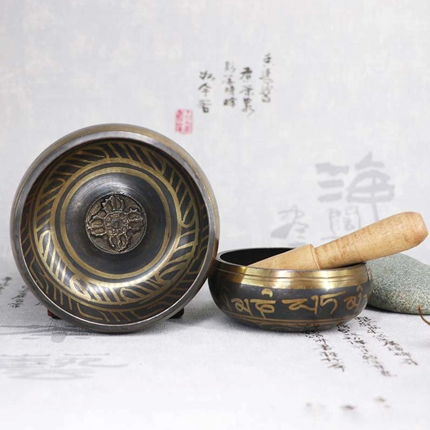Two sizes of Buddhist meditation singing bowls with golden Sanskrit engravings displayed in top-down flat lay, featuring wooden striker resting in smaller bowl