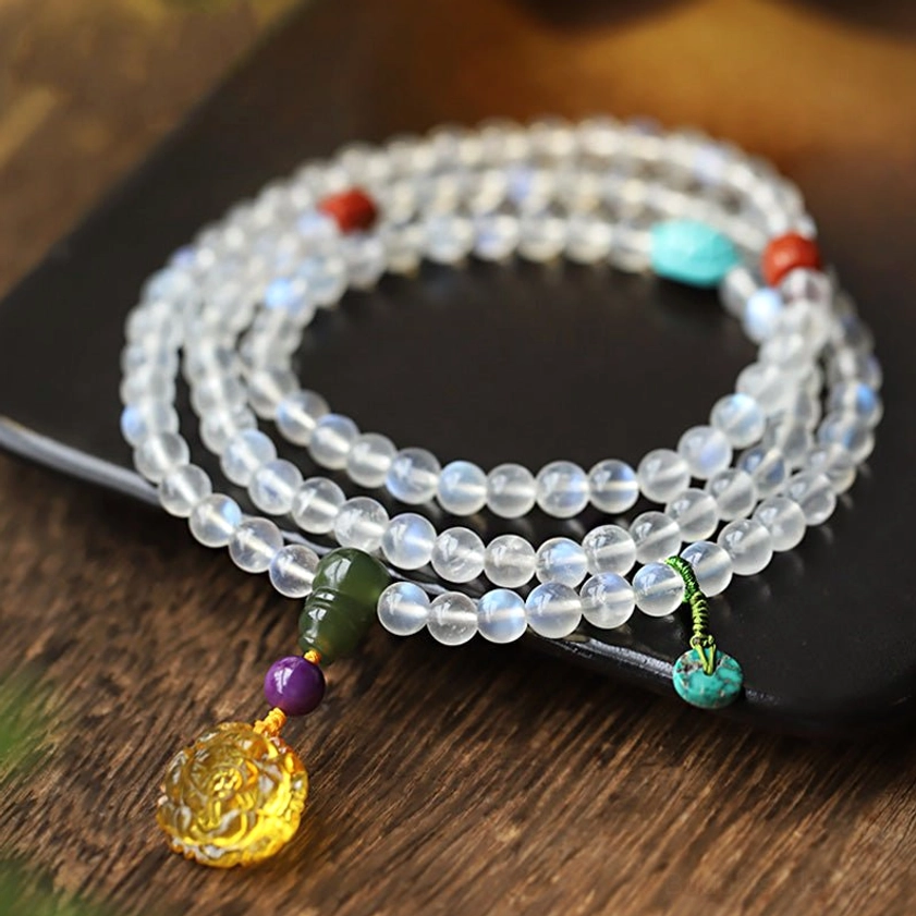 108 moonstone mala beads bracelet with carved amber lotus pendant, displayed on black tray with wooden table background
