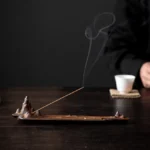 Brown ceramic mountain incense burner with lit incense stick on dark wooden tea table, showing realistic smoke rising and natural wood-grain texture detail