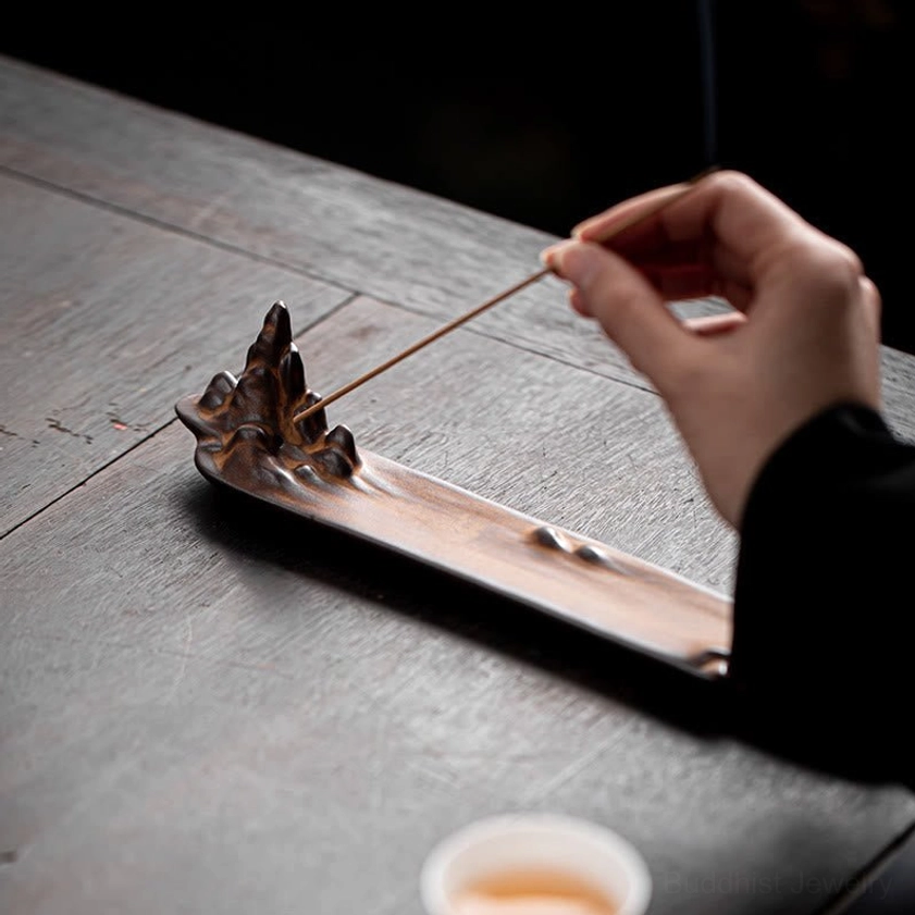 Eye-level side view of mountain ceramic incense holder on grey wooden table with hand positioning incense stick into burner slot