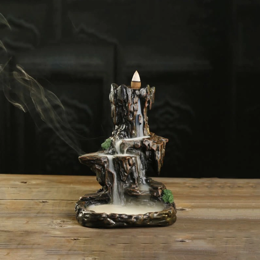 Lit Buddhist mountain waterfall incense burner with rising smoke, dark brown handcrafted resin, front angle view