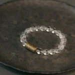 Multi-crystal protection bracelet with clear quartz and rutilated quartz beads on dark metal tray, flat lay overhead view