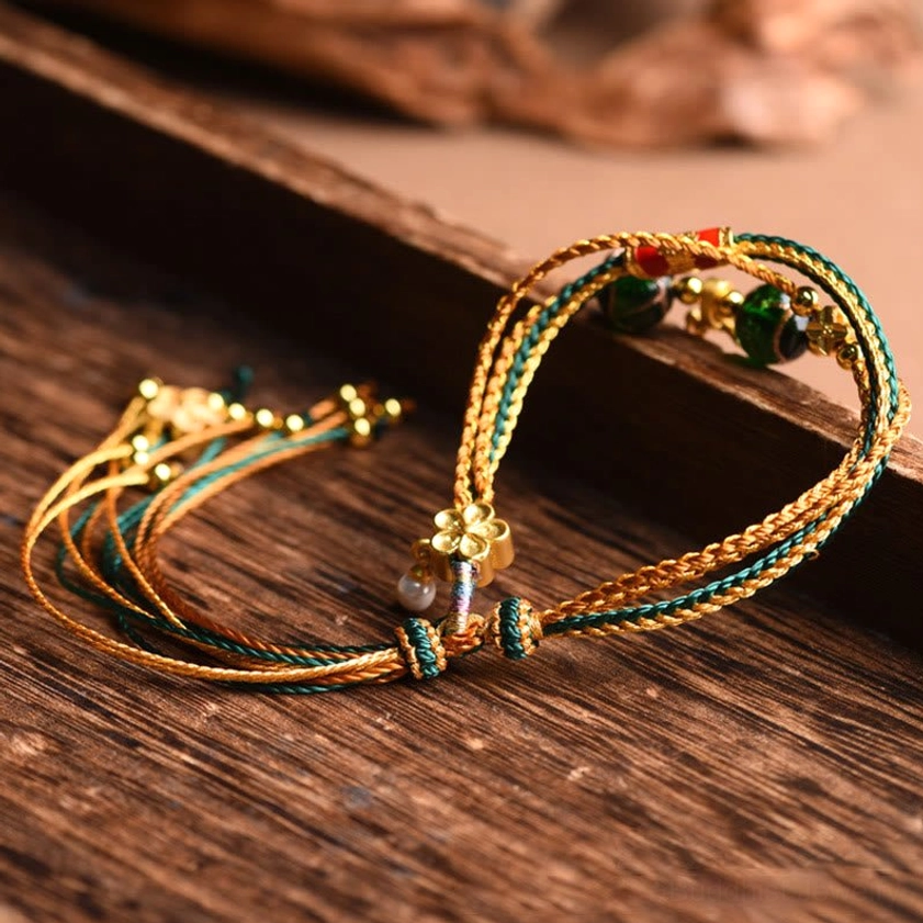 Multi-layered liuli glass bead bracelet unfolded showing gold-teal cords, dark green sand-blown glass beads, and metal flower accents