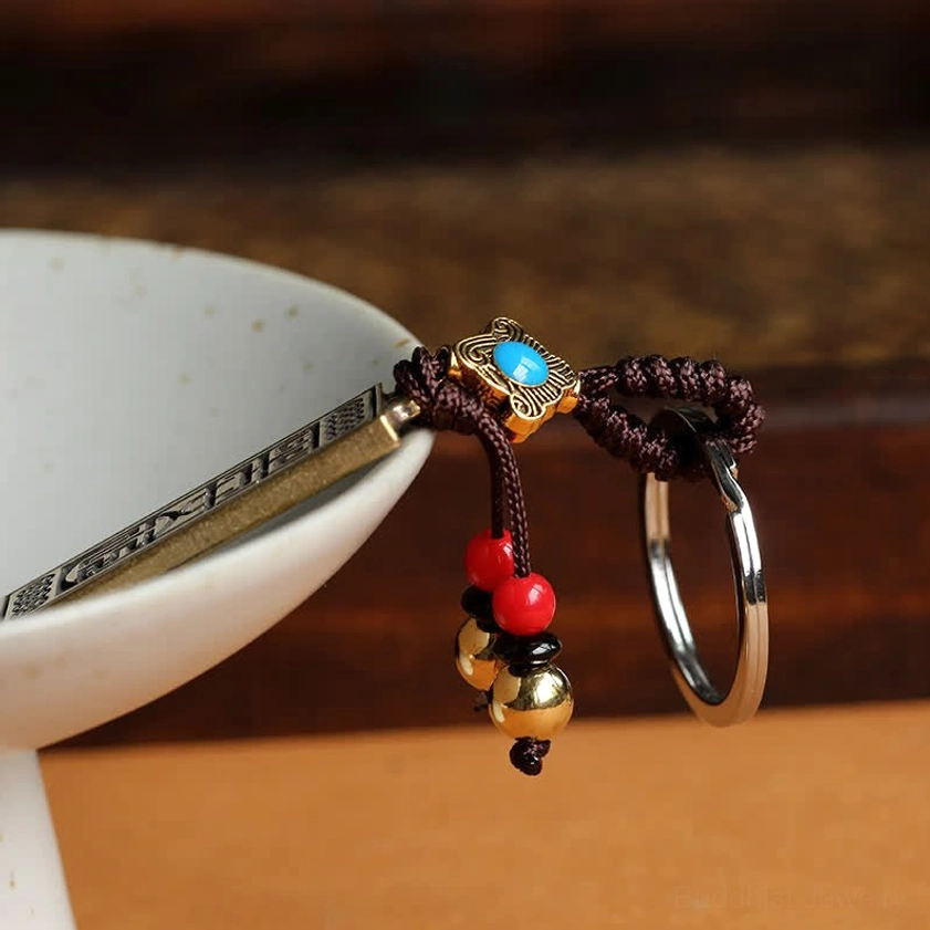 Low-angle side view of Buddhist keychain with long metal tag and blue enamel decoration beside white ceramic bowl