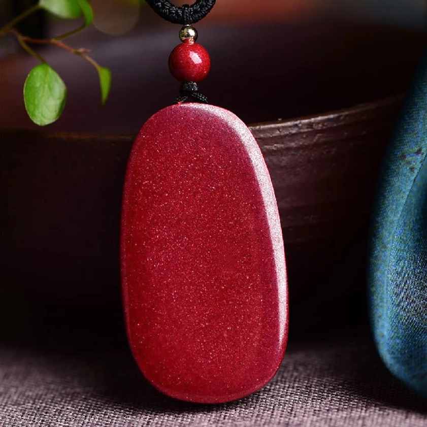 Solid natural cinnabar oval pendant necklace with smooth wine-red surface displayed on ceramic tabletop with botanical accents