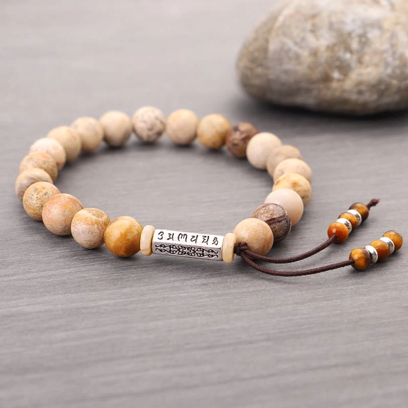 Weathered stone Om Mani Padme Hum bracelet with natural mixed tone beads and brown cord tassel on grey backdrop