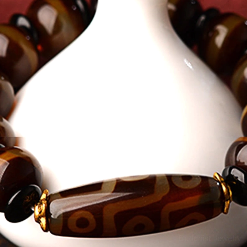 Nine-eye dzi bead agate bracelet close-up showing glossy brown striped agate texture and gold connectors with white ceramic vase backdrop