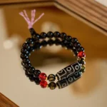 Nine-eye Dzi bead black onyx bracelet with Om Mani Padme Hum spacers and pink tassel, angled view on warm wooden surface