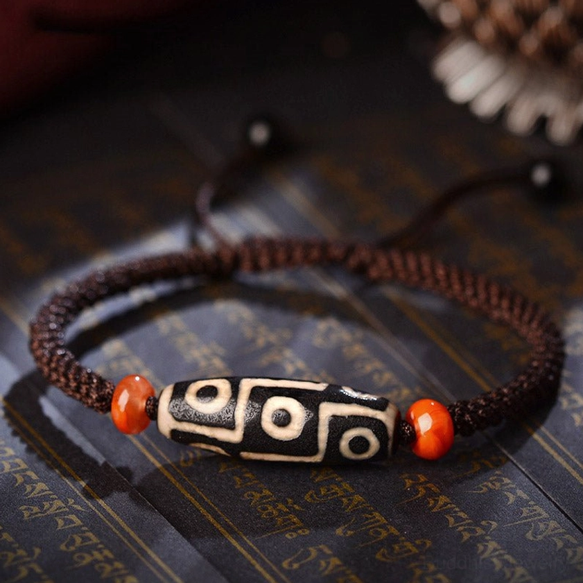 Nine-Eye Dzi Bead bracelet with brown braided cord and red agate spacers displayed on Buddhist scripture background