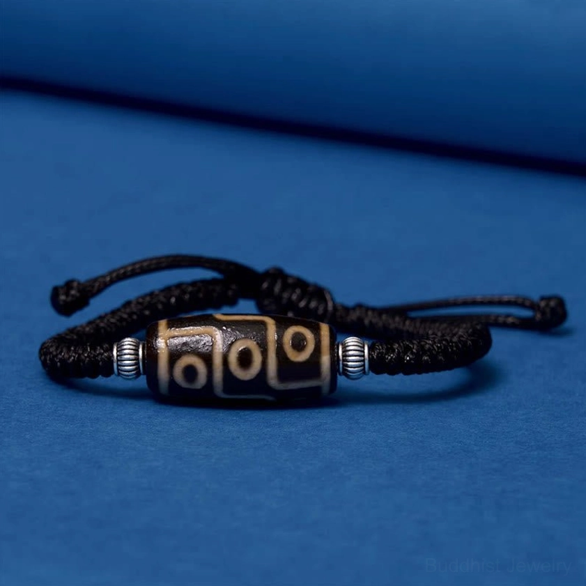 Nine-eye dzi bead bracelet side flat lay on blue background highlighting adjustable black braided rope and metal components