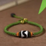Nine-eye dzi bead green rope bracelet with adjustable sliding knot, three-quarter front view showing black-white agate bead and braided cord