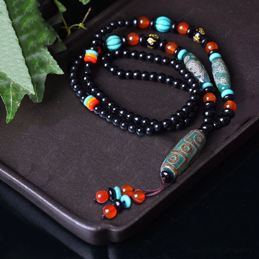 Nine-eye Dzi bead Buddhist necklace coiled flat lay view on dark display tray with decorative green leaves, showing complete beaded chain and traditional tassel design