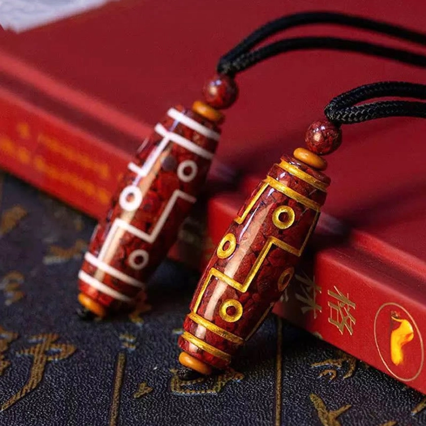 Two styles of nine-eye dzi bead Buddhist necklace pendants side by side, one with gold motifs and one with white motifs on black cords, displayed on vintage patterned desk