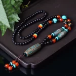 Nine-eye Dzi bead necklace with dragon pattern and Om Mani Padme Hum mantra pendant displayed on dark tray with green leaf styling, showing traditional beaded chain and tassel design