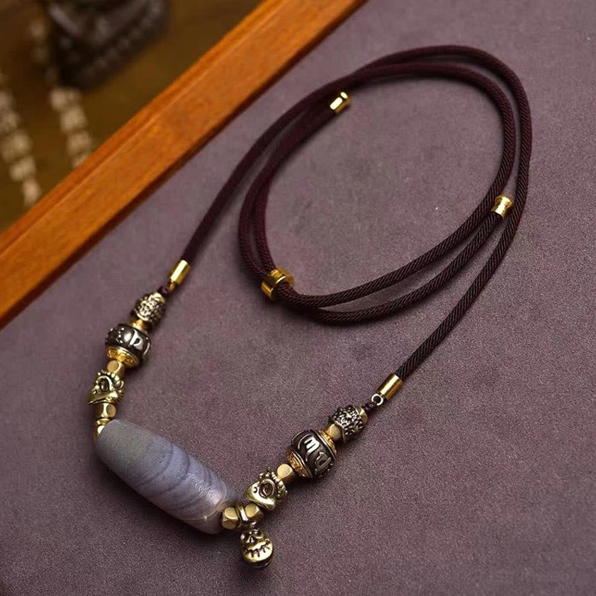 Nine-Eye Dzi Bead Om Mani Padme Hum Buddhist protection necklace flat lay with woven cord and metal mantra beads on dark textile