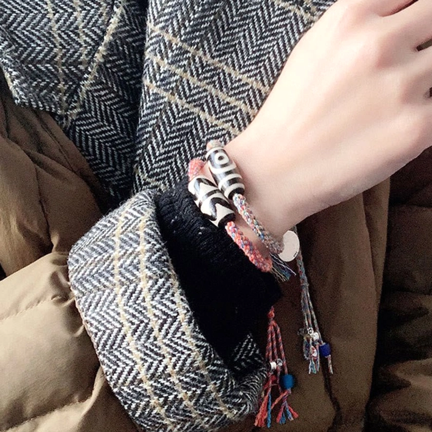Handcrafted Nine-Eye Dzi Bead protection bracelet on wrist featuring black-white dzi bead, silver charms, colorful tassel pendants, and braided cord