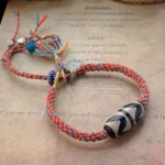Nine-Eye Dzi Bead Sterling Silver Om Mani Padme Hum red braided protection bracelet with silver charm and colored tassels on vintage paper