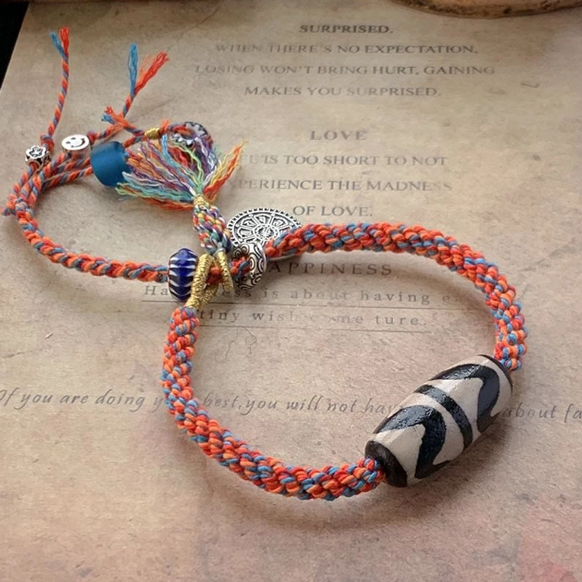 Nine-Eye Dzi Bead Sterling Silver Om Mani Padme Hum red braided protection bracelet with silver charm and colored tassels on vintage paper