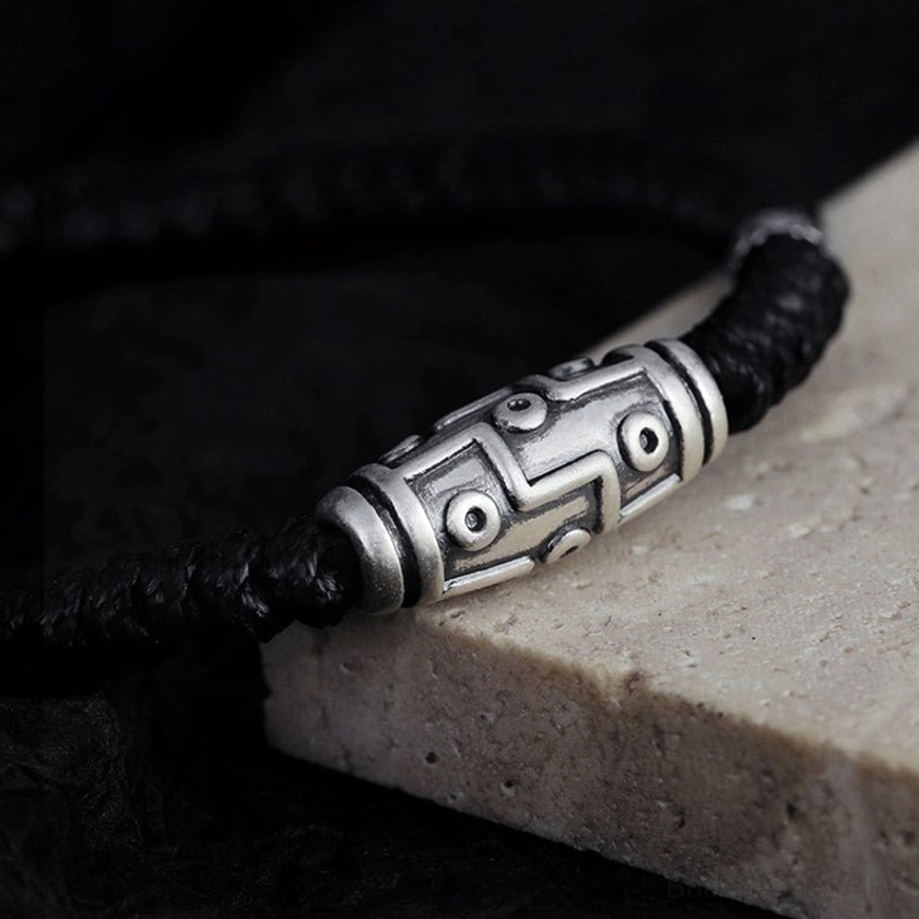 Low side view of 999 sterling silver nine-eye dzi bead bracelet with black woven cord on stone base with dark backdrop