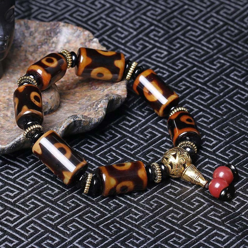 Nine-eye dzi bead protection bracelet on stone coaster with decorative fabric backdrop, showing polished stone beads and matching hardware
