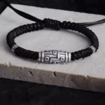 Overhead view of 999 sterling silver nine-eye dzi bead bracelet with black woven cord on light stone base, showing intricate dzi pattern