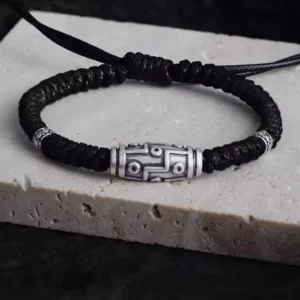 Overhead view of 999 sterling silver nine-eye dzi bead bracelet with black woven cord on light stone base, showing intricate dzi pattern
