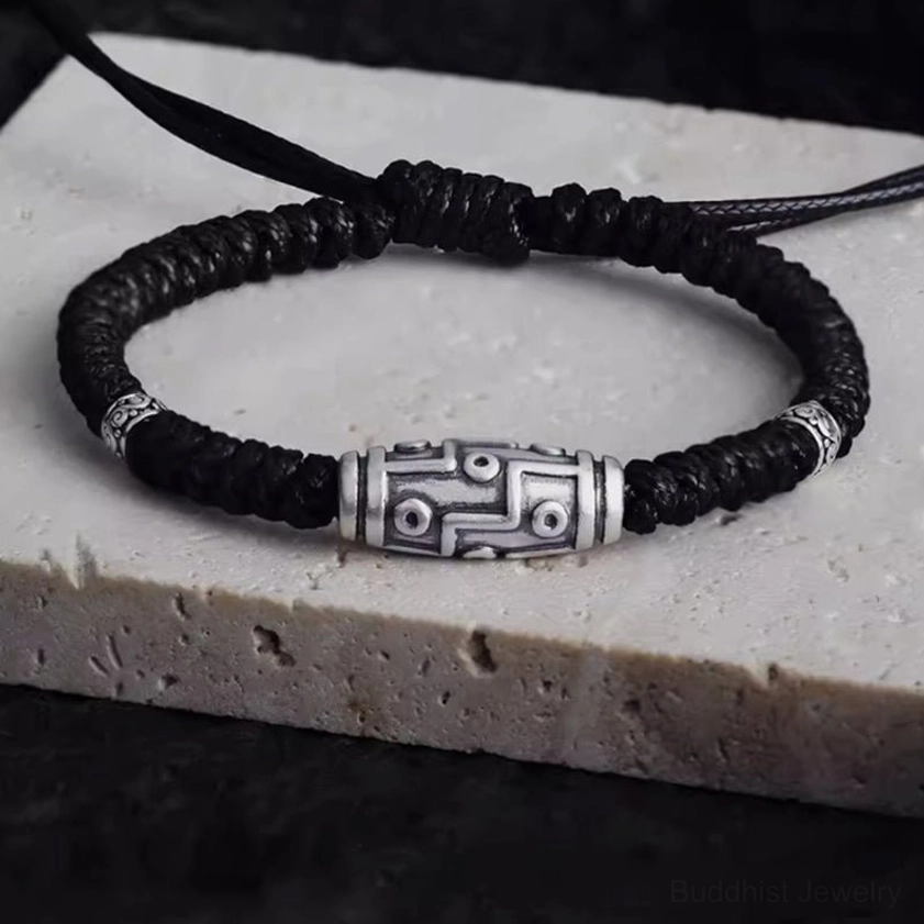 Overhead view of 999 sterling silver nine-eye dzi bead bracelet with black woven cord on light stone base, showing intricate dzi pattern