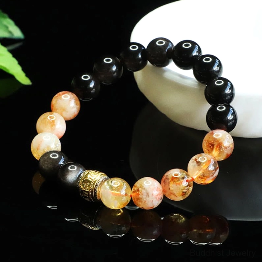 Buddhist healing bracelet featuring silver sheen obsidian and citrine gemstone beads with gold metal accents, top-down view on black reflective surface