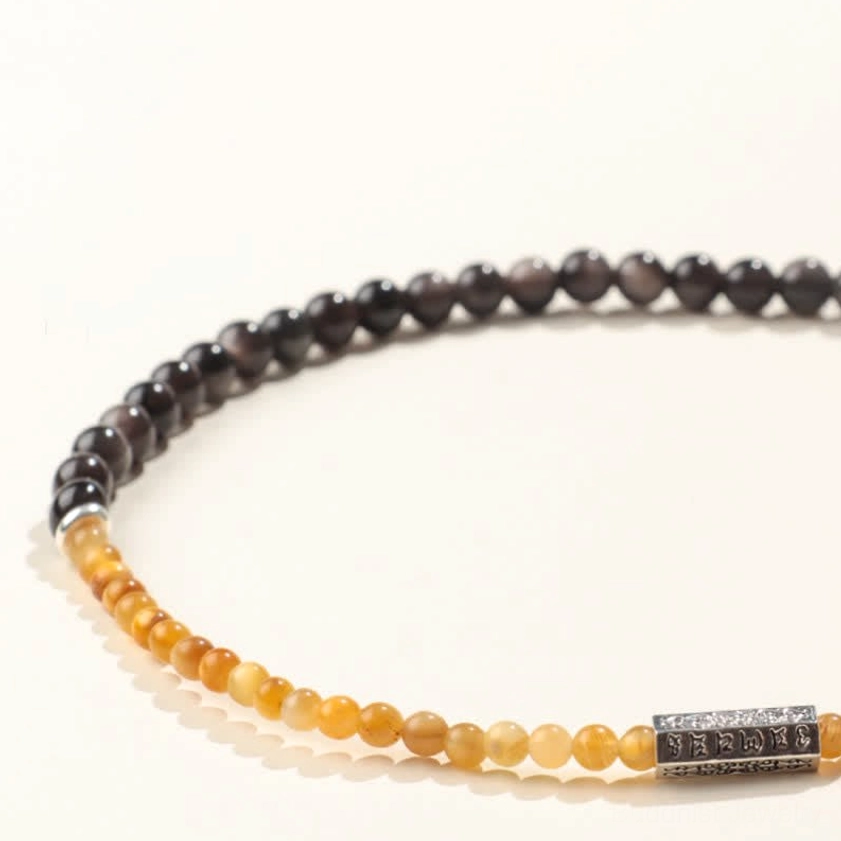 Side low-angle view of silver sheen obsidian double wrap bracelet showing gradient yellow-brown beads, dark stone, and engraved silver charm with depth perspective