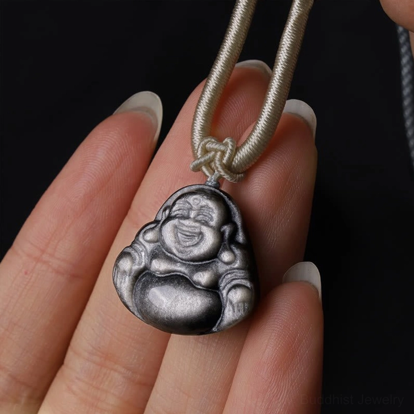 Polished silver sheen obsidian Buddhist Laughing Buddha pendant necklace held on fingertip, showing lustrous finish and beige braided cord