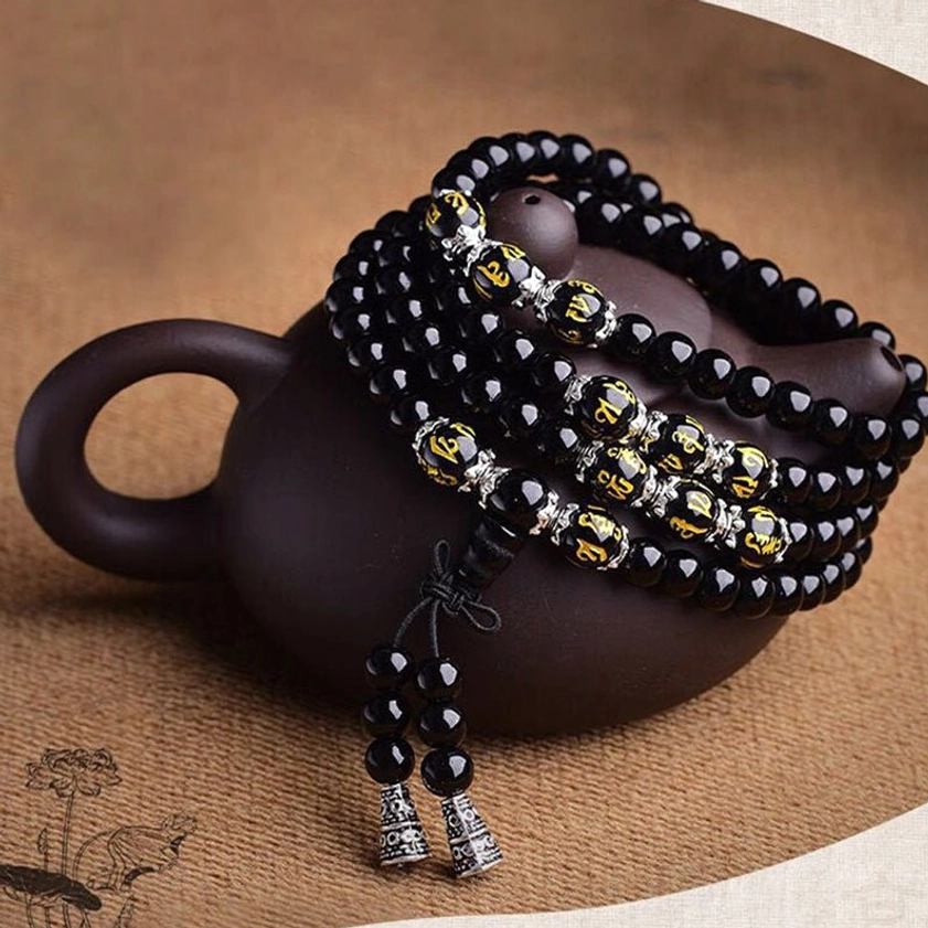 Black Obsidian Buddhist prayer mala bracelet with golden word beads and double-pendant tassel displayed on dark clay teapot