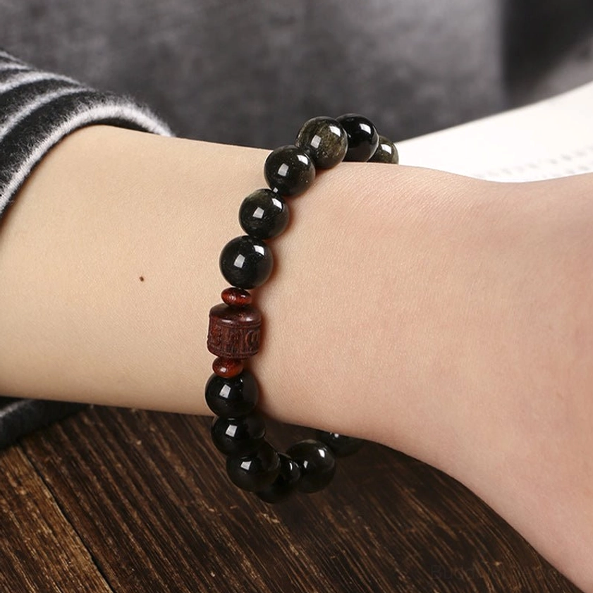 Om Mani Padme Hum obsidian bracelet worn on wrist on wooden table, showing polished black beads with golden sheen and reddish accent beads