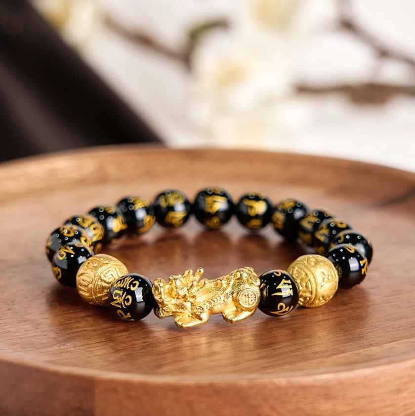 Pixiu obsidian bracelet flat lay on wooden tray with blurred floral background, classic black and gold color composition