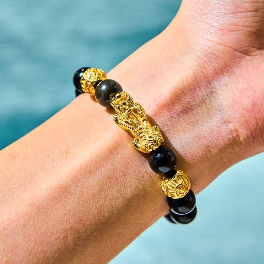 Buddhist Pixiu protection bracelet worn on wrist, side view displaying black obsidian beads with golden Pixiu charm and spacer beads against light blue background