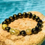 Black obsidian Pixiu wealth bracelet with Om Mani Padme Hum engraved beads and gold Pixiu charm, overhead view on turquoise water stone background