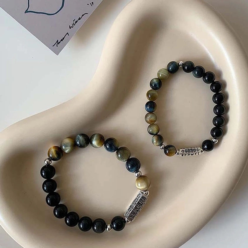 Two black obsidian tiger eye beaded bracelets separated on curved ceramic tray showing individual design with metal charm details