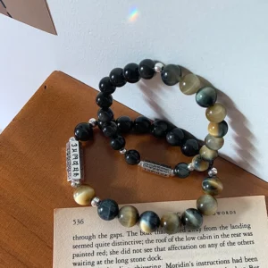 Black obsidian tiger eye Om Mani Padme Hum bracelet with engraved metal charm beads displayed on wooden surface and open book, flat lay Buddhist jewelry