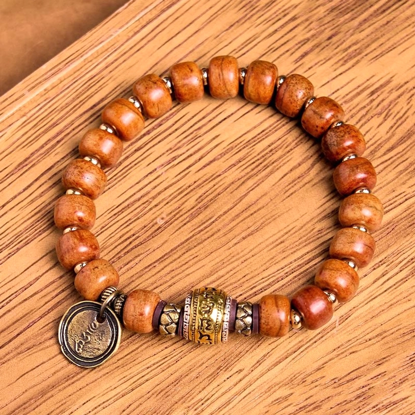 Top-down view of camel bone OM luck bracelet on wood grain background, showing tan barrel beads and engraved round metal pendant with Sanskrit symbol