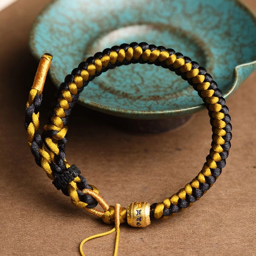 Buddhist string bracelet with dragon scale rope pattern and metal mantra bead, displayed beside turquoise ceramic bowl