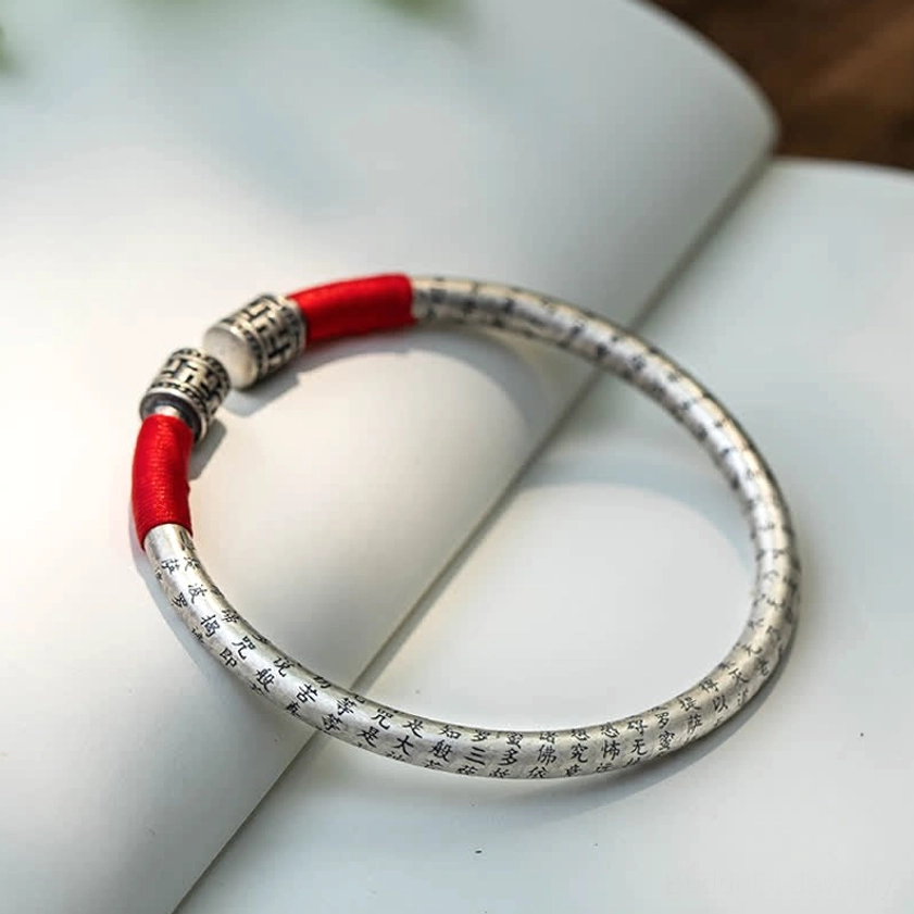 Buddhist Om Mani Padme Hum silver bangle with engraved text and red silk wrap, flat lay on spiritual book background