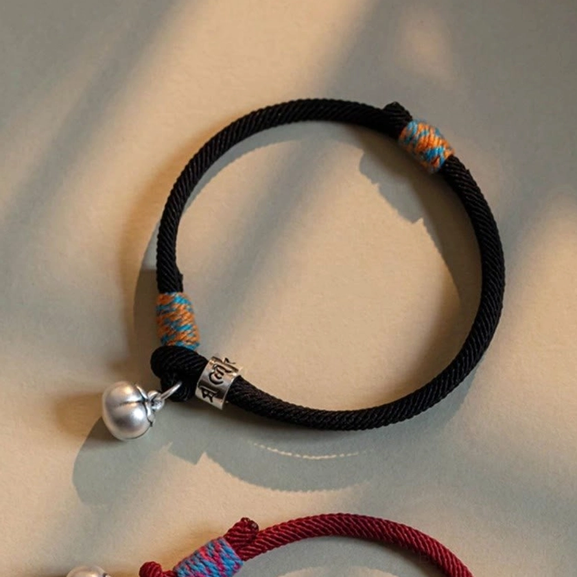 Close-up side view of black braided Buddhist bracelet showing engraved mantra bead and silver bell charm