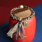 Om Mani Padme Hum handmade Buddhist bracelet with blue beige black woven cord and copper charm, displayed on decorative background