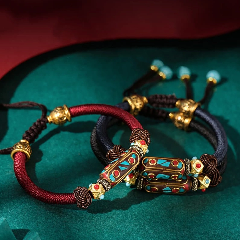 Close-up top view of Buddhist mantra bracelet showing red and dark blue braided ropes with ornate enamel charm and antique gold metal accessories on velvet surface